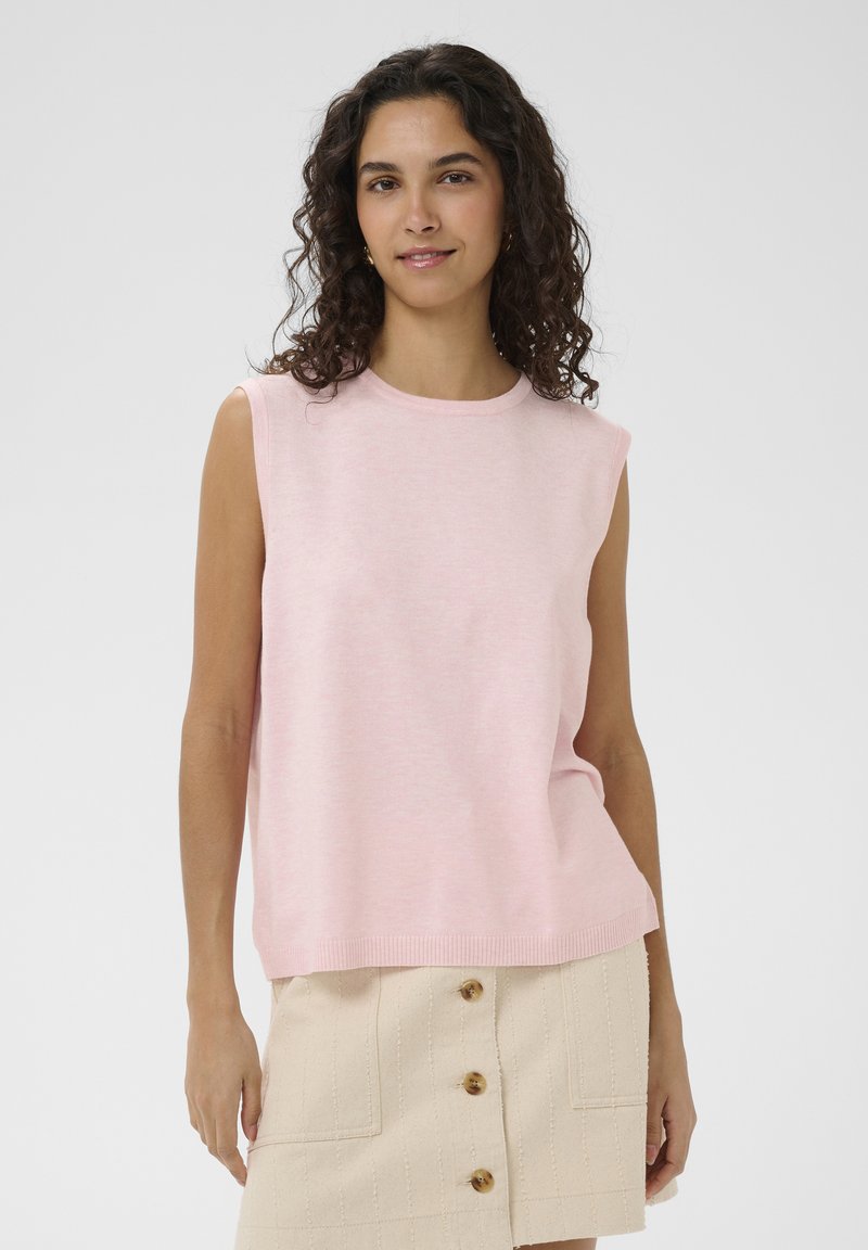 Femme aux cheveux bouclés et foncés, portant un haut sans manches rose clair et une jupe beige avec des boutons sur le devant, sur un fond uni.