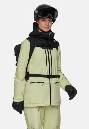 Geïsoleerde skijas in lichtgroen, met een rits aan de voorkant, zwarte accenten en een afneembare capuchon. Gecombineerd met zwarte handschoenen en een helm.