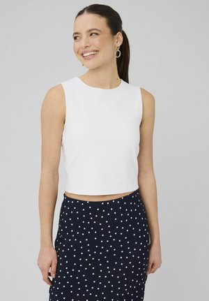 Femme souriante avec une queue de cheval foncée, portant un crop top blanc sans manches et une jupe bleu marine à pois blancs, debout devant un fond clair uni.