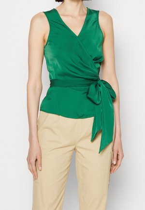 Vrouw draagt een groene mouwloze wikkeltop met strik in de taille en beige hoge taillebroek, staand tegen een effen achtergrond.