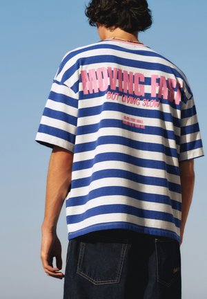 Persoon met krullend haar die een blauw-wit gestreept shirt draagt met roze tekst "MOVING FAST BUT LIVING SLOW" op de rug tegen een heldere lucht.