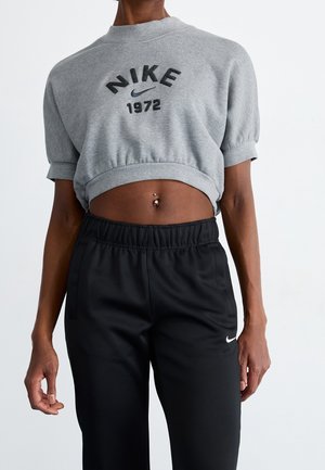 Personne portant un sweat-shirt Nike 1972 gris court et un pantalon Nike noir, montrant un piercing au nombril sur un fond uni.