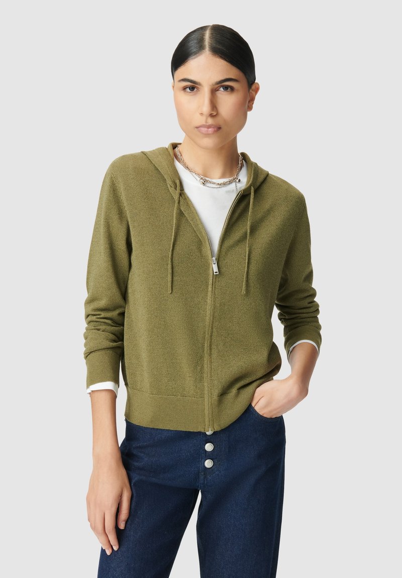 Junge Frau mit dunklem Haar in einem Mittelscheitel trägt einen olivgrünen Reißverschluss-Hoodie, ein weißes Shirt, mehrlagige silberne Halsketten und dunkelblaue, geknöpfte Jeans.