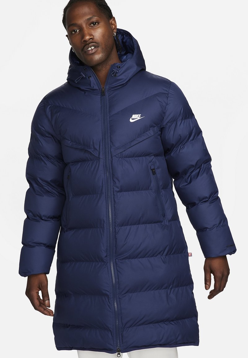 Marineblauer Daunenmantel mit Kapuze, diagonal geschnittenen Nähten, mit Reißverschlusstaschen und einem weißen Nike-Logo auf der Brust.