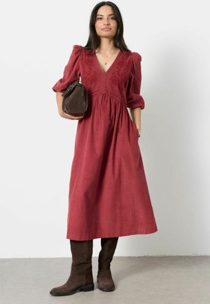 Woman in a long red dress with puffed sleeves, holding a brown handbag, wearing dark brown knee-high boots, standing on gray floor.