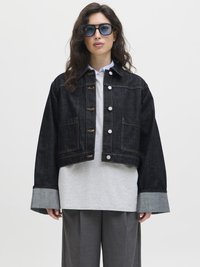 Black denim jacket with a cropped design, silver hardware, and large pockets, layered over a gray sweatshirt and wide gray trousers.