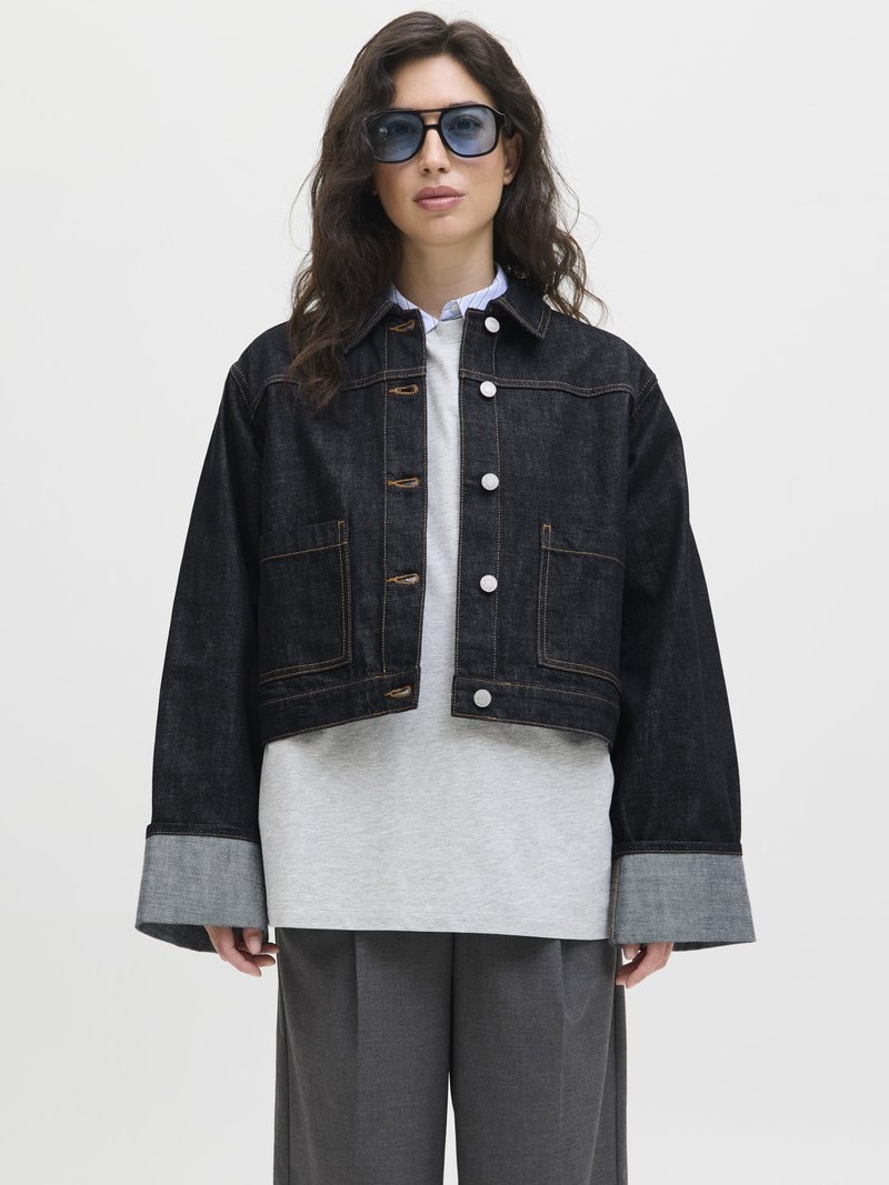 Black denim jacket with a cropped design, silver hardware, and large pockets, layered over a gray sweatshirt and wide gray trousers.
