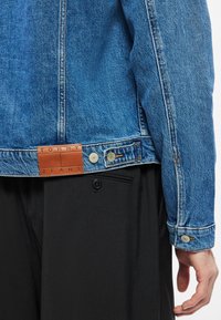 Világoskék farmerdzseki, bőr címkével, rajta a "TOMMY JEANS" felirattal. Fém gombokkal és kontrasztos varrási részletekkel rendelkezik. Alatta fekete nadrág látható.