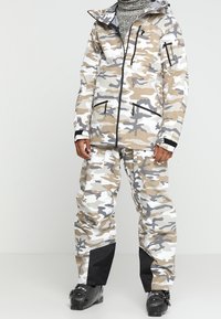 Kamouflage-mönstrad ytterkläder i beige, grå och vit. Jackan har huva, dragkedjefickor och kontrasterande svarta detaljer.