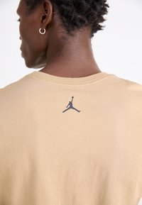 Béžové bavlněné tričko s malým černým logem Jordan na horní části zad. Materiál vypadá jemně a má hladkou strukturu.