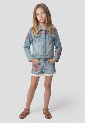 Niña joven de pie con las manos en los bolsillos, vistiendo una chaqueta ligera de mezclilla y shorts con bordado floral, y sandalias marrones.