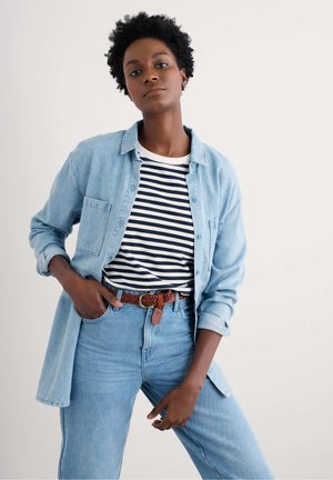 Camisa de mezclilla azul claro sobre una camiseta de rayas en blanco y negro, combinada con jeans azul claro y un cinturón trenzado marrón.