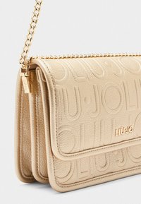 Sac bandoulière en cuir texturé beige avec un logo en relief et une bandoulière en chaîne dorée. Il dispose d'une fermeture à rabat et d'une forme structurée.