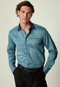 Cămașă teal cu butoni, cu un model punctat, guler navy contrastant și un singur buzunar la piept. Purtată cu pantaloni negri și o curea.