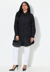 Tunic nera ricamata con ritagli floreali, maniche lunghe e chiusura a bottoni, abbinata a pantaloni bianchi aderenti e tacchi alti neri.