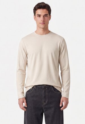Junger Mann mit kurzen dunklen Haaren, der ein beiges Langarmhemd und dunkle Jeans trägt und vor einem schlichten weißen Hintergrund steht.