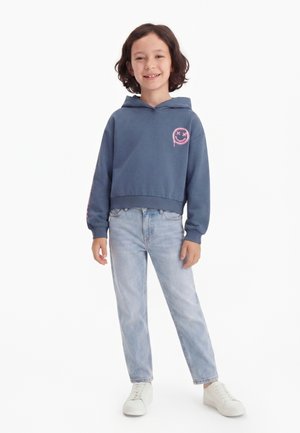 Enfant aux cheveux foncés mi-longs portant un sweat à capuche bleu avec un smiley rose, un jean bleu clair et des baskets blanches, debout sur un fond blanc.
