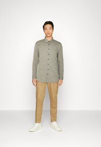 Camisa de botones de manga larga gris con cuello estructurado, combinada con pantalones marrón claro y zapatillas blancas. Textura lisa, diseño casual.