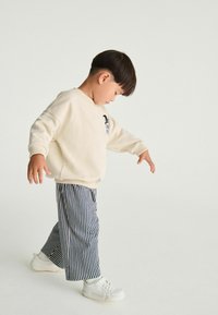Beige sweatshirt met geribbelde cuffs, marineblauwe en witte gestreepte wijde broek, en witte sneakers. Kind loopt met het hoofd naar beneden.