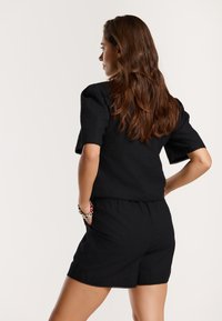 Compleu negru format din două piese, incluzând un top cu mânecă scurtă și pantaloni scurți. Material texturat cu o margine ondulată și buzunare laterale.