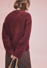 Burgundowa dziana bluza z prążkowanymi mankietami i dołem, luźny krój, teksturowana tkanina, noszona z brązowymi spodniami i brązową torbą, widok z tyłu.