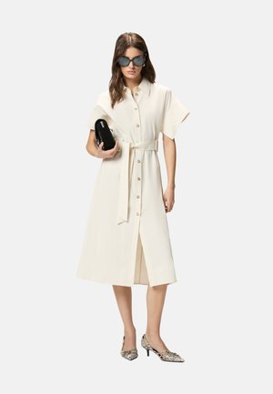 Donna che indossa un vestito midi crema con bottoni e cintura in vita, tacchi con motivo, grandi occhiali da sole e tiene in mano una pochette nera.