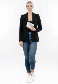 Blazer noir ajusté, haut noir cintré, jeans bleus taille haute et baskets blanches. Tenant un portefeuille blanc avec des accents argentés.