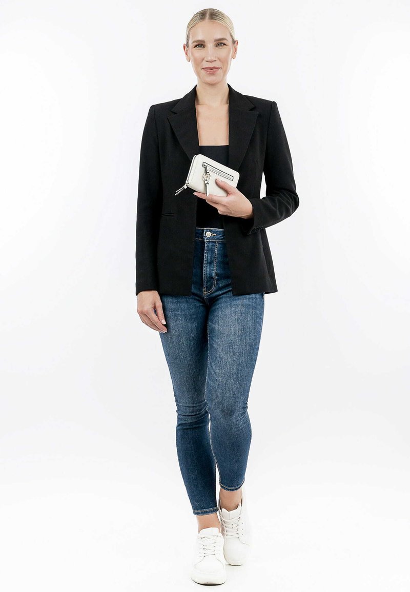 Blazer noir ajusté, haut noir cintré, jeans bleus taille haute et baskets blanches. Tenant un portefeuille blanc avec des accents argentés.