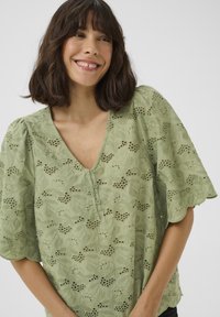 Blusa verde con scollo a V, maniche corte gonfie, ricami floreali e chiusura con bottoni caratterizzata da motivi a intaglio. Tessuto leggero e texture morbida.