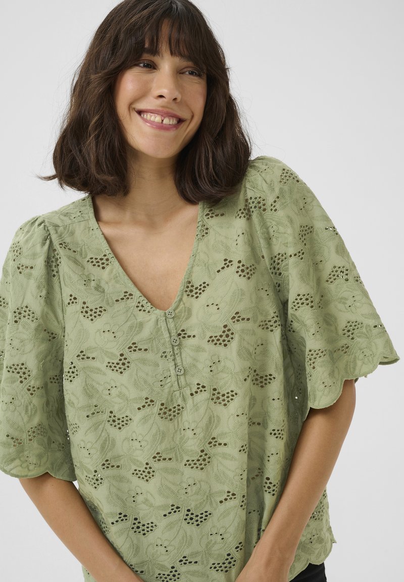 Blusa verde con scollo a V, maniche corte gonfie, ricami floreali e chiusura con bottoni caratterizzata da motivi a intaglio. Tessuto leggero e texture morbida.