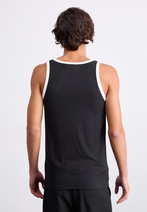 Mann mit kurzen lockigen Haaren, der ein schwarzes ärmelloses Shirt mit weißem Besatz trägt, steht mit dem Rücken zur Kamera vor einem schlichten weißen Hintergrund.
