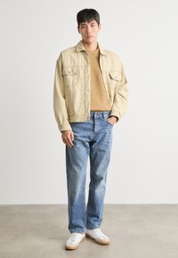 Chaqueta de mezclilla color beige claro con dos bolsillos en el pecho, llevada sobre un suéter marrón, combinada con jeans azul claro y zapatillas blancas con suelas color beis.