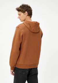 Bruine hoodie van zachte stof, met een kangoeroezak, ribgebreide boorden en onderrand, gedragen met donkere jeans. Glad textuur en casual ontwerp.