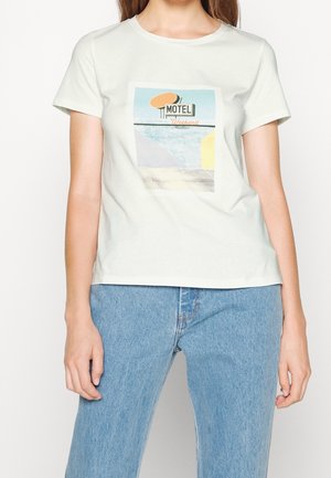 Vrouw draagt een wit T-shirt met korte mouwen en een grafische afbeelding van een motelteken in blauw, geel en oranje, gecombineerd met lichtblauwe spijkerbroek.