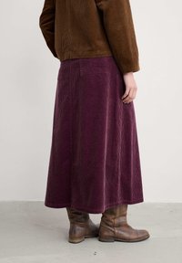 Brūna korduroja jaka, apvienota ar garu, tumši purpursarkanu korduroja svārku. Svārkiem ir teksturēta apdare un divas aizmugurējās kabatas. Brūni zābaki papildina apģērbu.