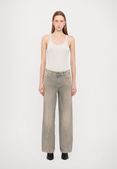 Hellgraues Tanktop, weite beige Jeans mit vorderen Taschen und verblassten Details, kombiniert mit schwarzen Absatzstiefeln, Model steht vor einem schlichten Hintergrund.