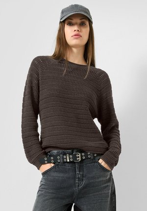 Frau mit langen Haaren, die eine graue Kappe, einen dunkelbraunen strukturierten Pullover, einen schwarzen Ösengürtel und ausgewaschene schwarze Jeans trägt und die Hände in den Taschen hat.