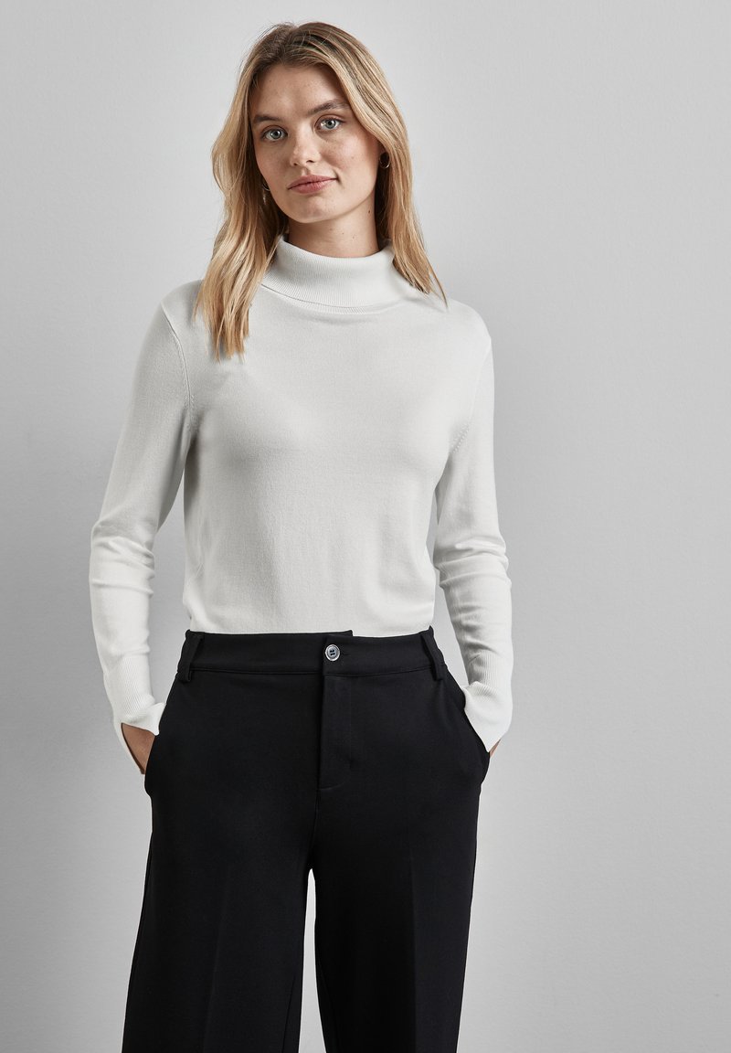 Weißer Rollkragenpullover mit langen Ärmeln, glatter Textur. Kombiniert mit hoch taillierten schwarzen Hosen mit Knopfverschluss und seitlichen Taschen.