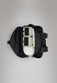 Mochila preta com uma abertura com fecho que mostra dois compartimentos cinza claro, fixos com correias pretas, forro suave e um detalhe de metal circular.