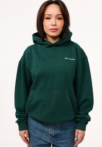 Dunkelgrüner Hoodie aus Baumwolle, mit einer Fronttasche, weiten Ärmeln und dem weißen "DAILY COTTON"-Logo auf der Brust.