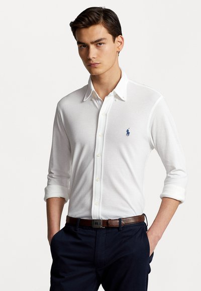 Weißes, langärmliges Hemd mit hochgekrempelten Ärmeln bis zum Ellbogen, das mit einem kleinen blauen Polo-Logo versehen ist. Kombiniert mit marineblauen Hosen und einem braunen Gürtel.