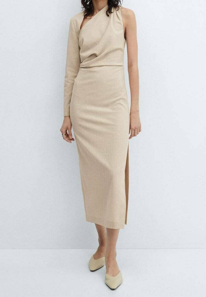 Femme portant une robe midi beige à une épaule avec une manche longue sur le bras gauche et des chaussures pointues beiges, debout contre un mur blanc.