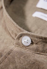 Gros plan d’un col de chemise en lin beige fixé par un bouton blanc à quatre trous.