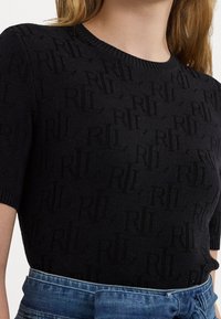 Černý pletený top s jemným texturovaným vzorem písmen, krátkými rukávy a kulatým výstřihem, kombinovaný s džíny v pase.