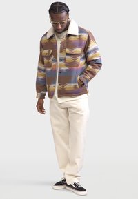 Giacca in misto lana con un motivo geometrico multicolore, collo in pelliccia shearling e due tasche sul petto, abbinata a pantaloni chiari e sneaker nere.