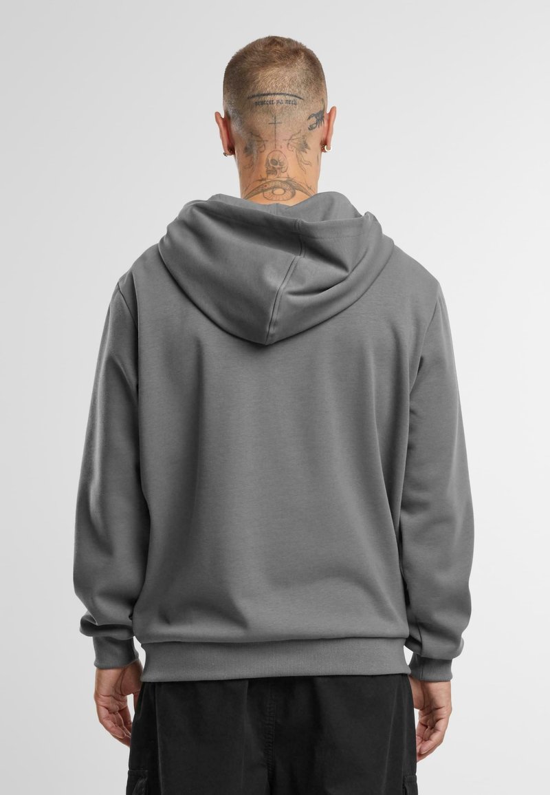 Personne aux cheveux courts et tatouages au cou, portant un sweat à capuche gris et un pantalon noir, tournée de dos sur un fond clair uni.