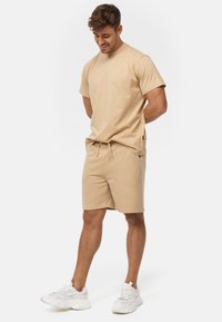 Ung mand, der kigger ned, iført beige t-shirt og shorts med hvide sneakers, stående mod en ensfarvet hvid baggrund.