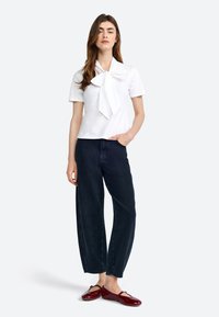 Weißes kurzärmeliges Oberteil mit einem großen Schleifendetail, kombiniert mit dunkelblauen weiten Jeans und glänzenden roten Slippern. Einfarbiger Hintergrund.