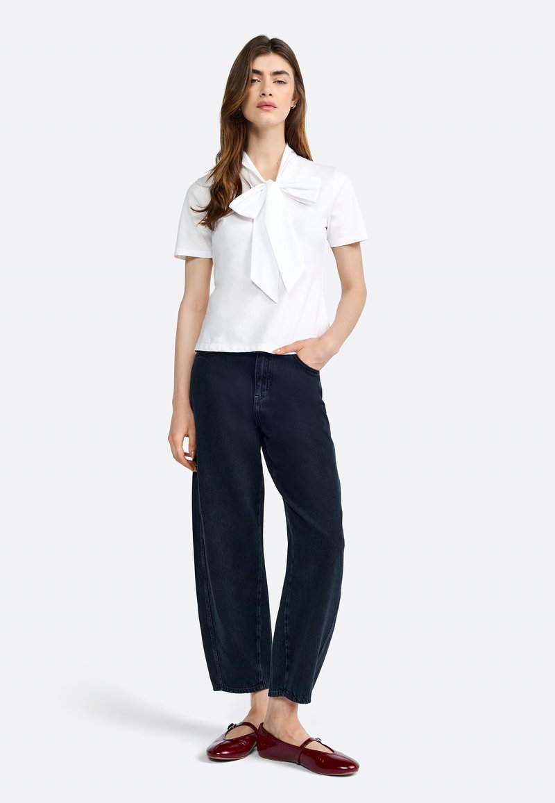 Weißes kurzärmeliges Oberteil mit einem großen Schleifendetail, kombiniert mit dunkelblauen weiten Jeans und glänzenden roten Slippern. Einfarbiger Hintergrund.