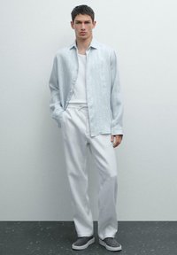 Chemise boutonnée en lin bleu clair sur un débardeur blanc, accompagnée d'un pantalon blanc ample et de baskets grises sur un sol sombre texturé.